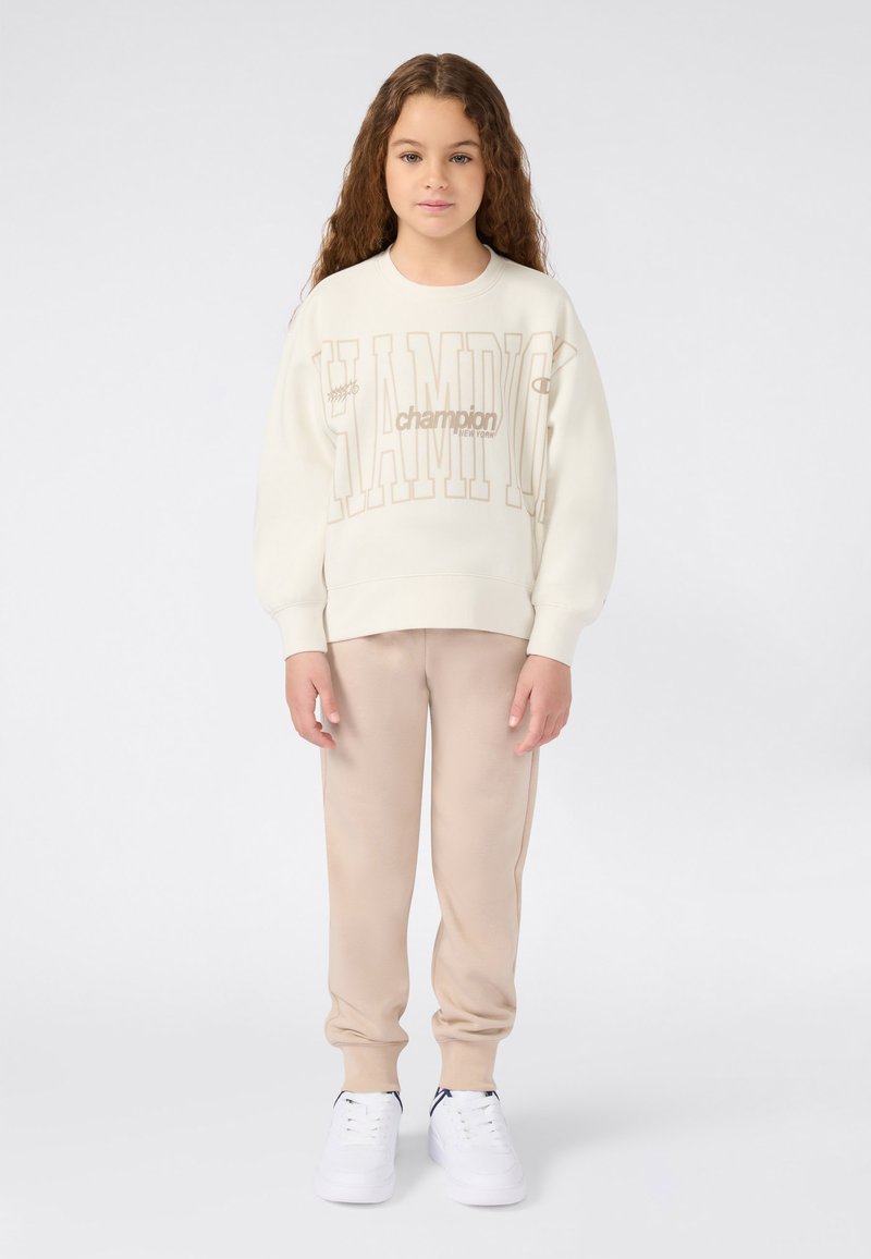 Niña pequeña con cabello rizado y largo, vistiendo pantalones de chándal beige, sudadera crema de Champion y zapatillas blancas, de pie contra un fondo simple.