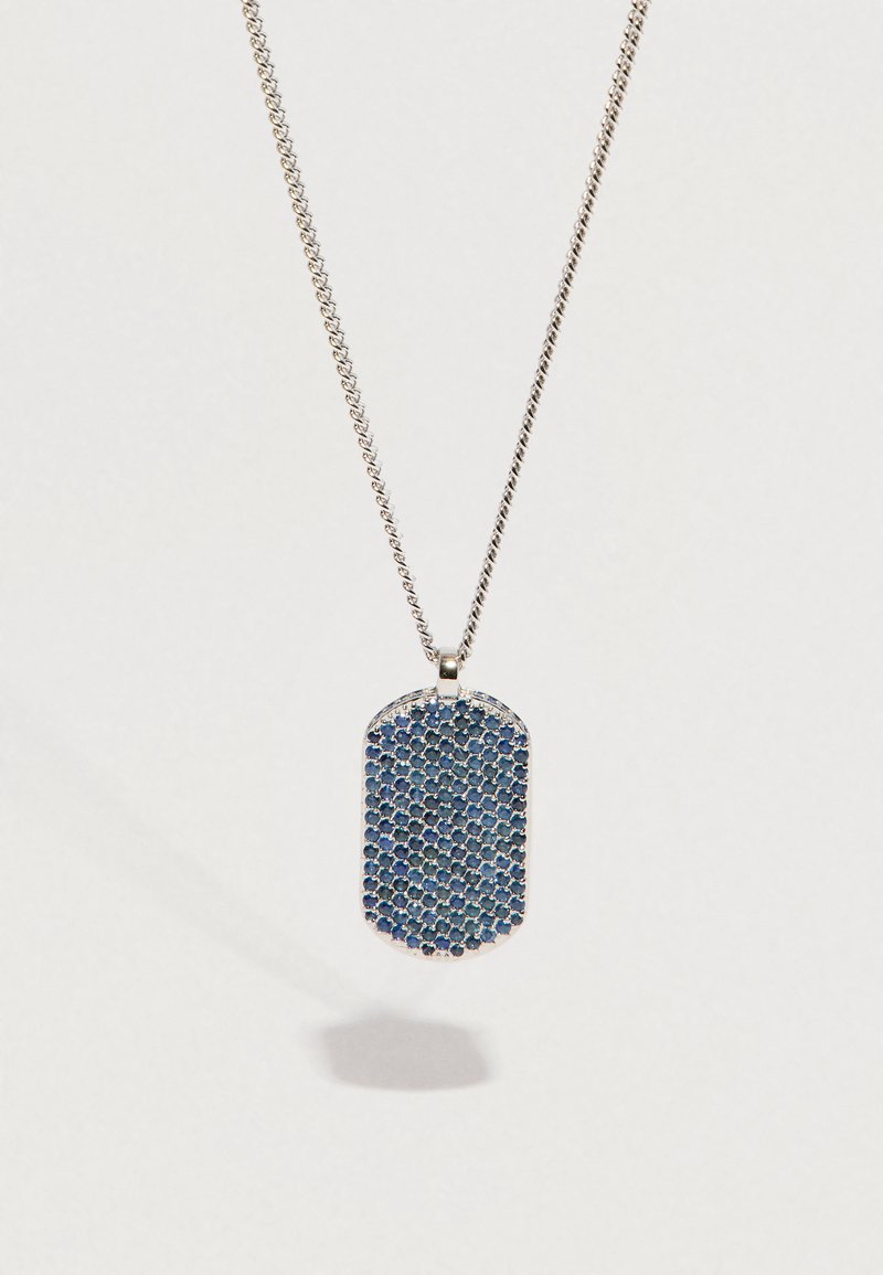 Stříbrný řetízek s obdélníkovým přívěskem vykládaným malými kulatými modrými drahokamy, vrhající stín na bílém pozadí.