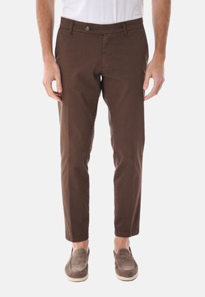 Uomo con pantaloni marroni a gamba dritta e scarpe marroni senza lacci, in piedi su uno sfondo chiaro e neutro.