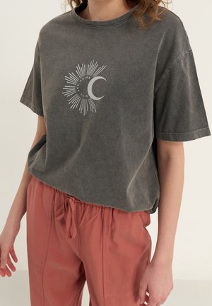 Woman wearing a faded black T-shirt with sun and crescent moon graphic and rust-colored drawstring pants, standing with hands relaxed.