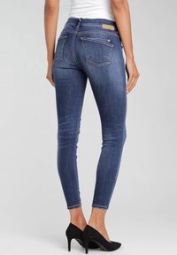 Vaqueros ajustados en denim azul oscuro con un ligero desgaste, diseño de cinco bolsillos y una etiqueta de cuero de marca en la cintura. Llevados con tacones negros.