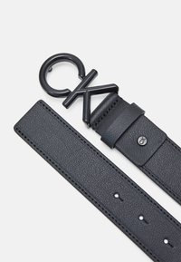 Cinturón de cuero negro con una hebilla circular que presenta el logo "CK" en relieve. Correa texturizada con múltiples agujeros para un ajuste regulable.