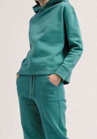 Ensemble pantalon de survêtement et sweat à capuche bleu sarcelle en matière douce. Présente une coupe décontractée, une taille avec cordon de serrage et un détail de logo subtil sur le pantalon.