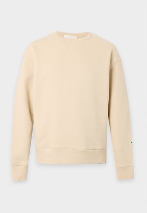 Hombre con sudadera beige encima de camisa azul clara con cuello, pantalones marrones holgados y zapatillas blancas, de pie con una mano en el bolsillo.