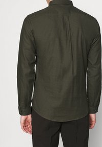 Camicia a maniche lunghe in verde scuro, tessuto testurizzato. Presenta un colletto con bottoni e un orlo curvo, con polsini con bottoni sulle maniche.