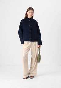 Navy Wolle-Mischung Jacke mit hohem Kragen und Frontknopfverschluss, kombiniert mit cremefarbenen, mit Pailletten versehenen, weit geschnittenen Hosen und einer grünen Handtasche.