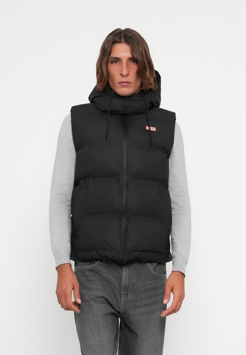 Melnā pūkaina veste ar kapuci, ar rāvējslēdzēju, auklu akcentiem un mazu sarkanu logo. Valkāta virs pelēka garām piedurknēm krekla.
