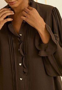 Donna che sistema una blusa marrone scuro con rouches e bottoni a forma di cuore, mostrando un primo piano delle mani e del scollatura.