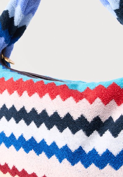 Πολύχρωμη τσάντα με ύφασμα σε σχέδιο ζιγκ ζαγκ και υφασμένη, βρόγχινη λαβή σε αποχρώσεις του μπλε, κόκκινου, λευκού και μαύρου.