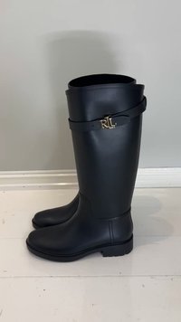 Botas altas de goma negra con textura suave, con un emblema dorado "RL" y un detalle de correa negra alrededor de la parte superior de la caña.
