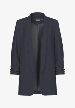 Vrouw met een marineblazer met opgestroopte mouwen, een zwarte top en een blauwe high-waisted jeans, staande tegen een effen achtergrond.