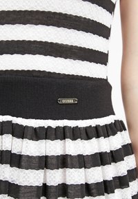 Robe à rayures noires et blanches avec une taille côtelée, un motif en dentelle et une petite plaque en métal portant l'inscription "GUESS". Design de jupe plissée.