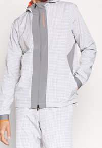 Veste légère avec un motif à carreaux gris et blanc, dotée d'une fermeture éclair frontale, d'une capuche ajustable et de poignets élastiques. Accent orange notable.