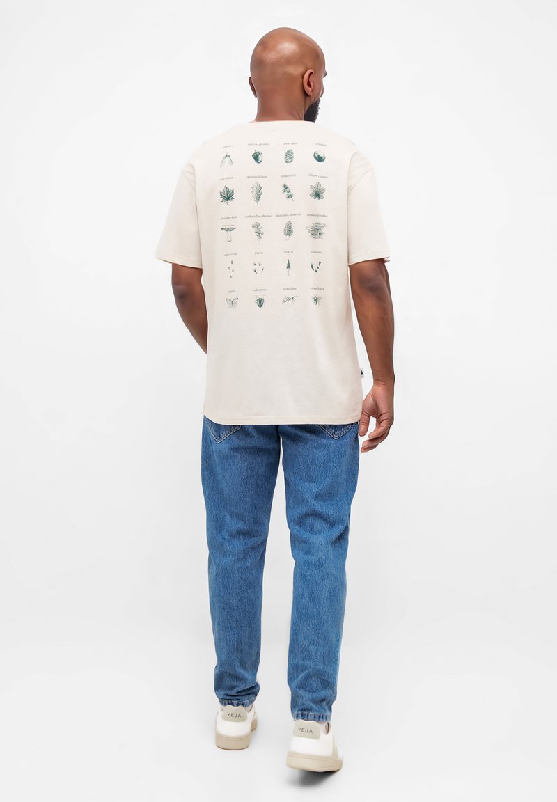 T-shirt léger beige à manches courtes avec des illustrations botaniques vertes au dos, associé à un jean bleu et des baskets blanches.