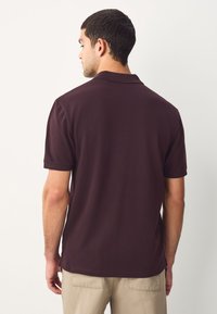 Tamsiai bordo spalvos trumpomis rankovėmis polo marškinėliai su klasikiniu apykakle, lygiu tekstūra ir tiesiu nugaros kirpimu, derinami su šviesiai smėlio spalvos kelnėmis.