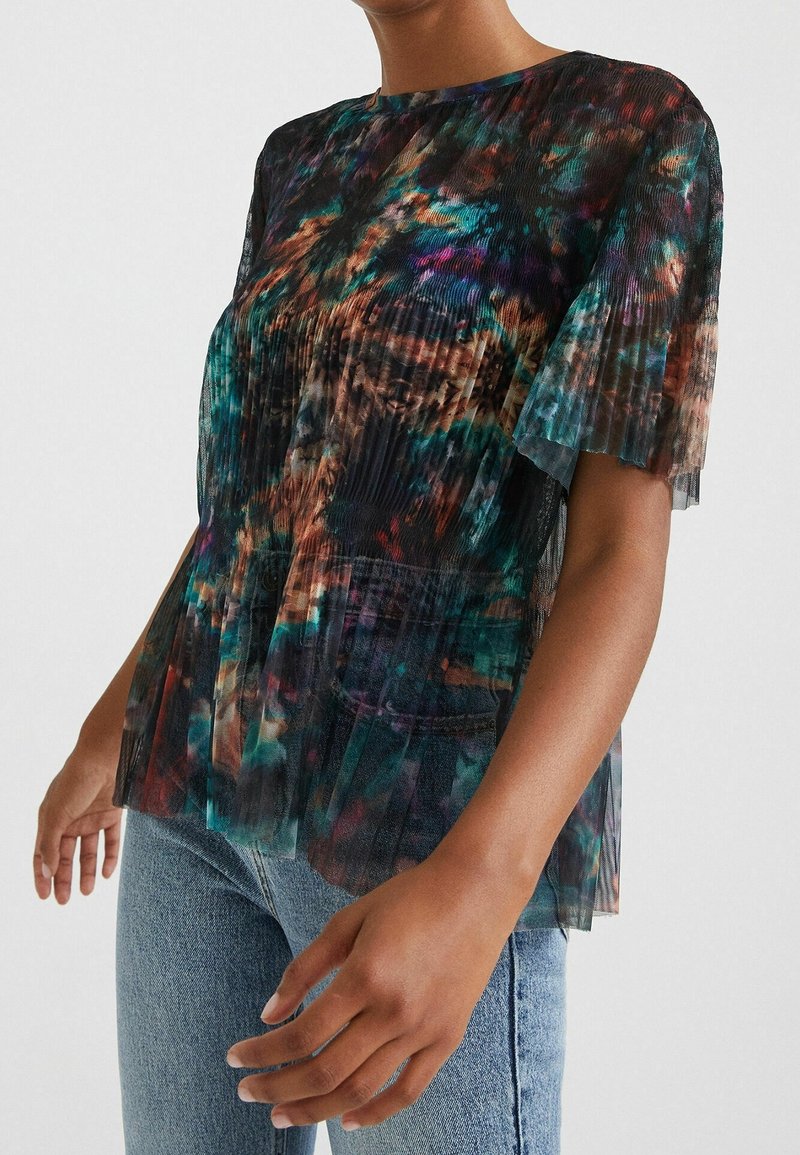 Personne portant un chemisier transparent plissé à manches courtes avec un motif abstrait multicolore et un jean en denim bleu clair.