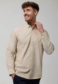 Camisa beige de manga larga con botones y cuello puntiagudo, que presenta una tapeta frontal, puños con botones y un sutil logo bordado en el pecho.