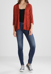 Mujer vistiendo un cárdigan estampado color óxido, camiseta negra, jeans ajustados azules y zapatillas bajas grises, de pie contra un fondo sencillo.