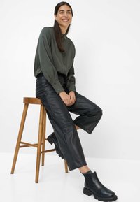 Sorte læder ankelstøvler, grøn skjorte og bukser i sort faux læder med bred ben. Model siddende på en træstol, smilende, mod en ensfarvet baggrund.
