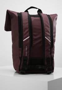 Sac à dos roll-top bordeaux avec des bretelles noires, des boucles ajustables et des accents réfléchissants. Fabriqué en tissu durable, il présente un logo minimal.