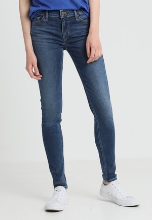 Jeans ajustados en denim azul oscuro, con un diseño clásico de cinco bolsillos, cremallera y cierre de botón, combinados con zapatillas blancas.