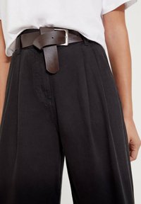 Brązowy skórzany pasek z kwadratową srebrną klamrą, noszony na wysokim stanie, luźnych czarnych spodniach. Teksturowany materiał z widocznymi bocznymi kieszeniami.