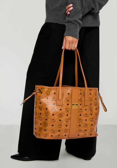 Donna in pantaloni neri che tiene una grande borsa tote MCM color beige con stampa nera del logo e doppi manici al suo fianco.