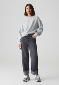 Grauer Sweatshirt mit überschnittenen Schultern, kombiniert mit dunklen Jeans, die an den Säumen gerollt sind. Das Model trägt graue Turnschuhe.