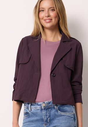 Junge Frau trägt eine dunkelviolette kurze Jacke über einem hellvioletten Top und blauen Jeans, lächelt mit den Händen in den Taschen.
