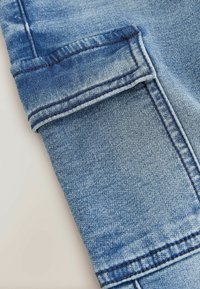 Secțiune pantaloni din denim, albastru deschis, cu tiv răsucit vizibil. Prezintă cusături contrastante și o textură fină cu un ușor luciu.