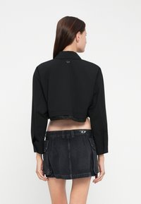 Korte zwarte jas met een gestructureerde kraag en lange mouwen, met een gegolfde zoom en een logo-accent op de achterkant. Gepaard met een donkerdenim rok.