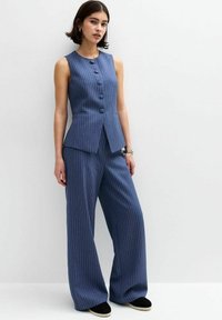 Ensemble comprenant un gilet à rayures bleues et un pantalon large. Le gilet présente des boutons et une coupe ajustée ; le tissu semble texturé. Les chaussures sont des espadrilles noires.
