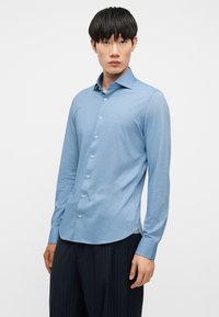 Νεαρός άνδρας που φοράει ανοιχτό μπλε πουκάμισο με κουμπιά και σκούρο παντελόνι με ρίγες, στέκεται μπροστά σε απλό λευκό φόντο.