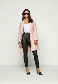 Lichtroze lange jas over een witte kanten top, gecombineerd met zwarte leren broeken en zwarte sandalen met hakken. Het model draagt zonnebrillen.