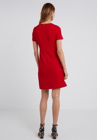 Robe rouge à manches courtes avec une coupe ajustée et une fermeture éclair au dos. Tissu texturé et ourlet évasé. Sandales à talons noirs cloutées visibles.