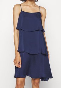 Marinblå lagerklänning med tunna band, som har en A-linjeformad silhuett och slät, glänsande tyg. Fyndiga volanger i fållen ger extra detalj.