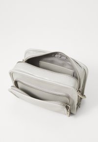 Šedá taška z umělé kůže s několika zipovými přihrádkami, hladkým povrchem a stříbrným kováním. Uvnitř se nachází přihrádka na karty.