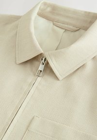 Camisa beige con cremallera, de tejido texturizado, que cuenta con un cuello puntiagudo y un solo bolsillo en el pecho, adornada con un hardware de cremallera plateada.