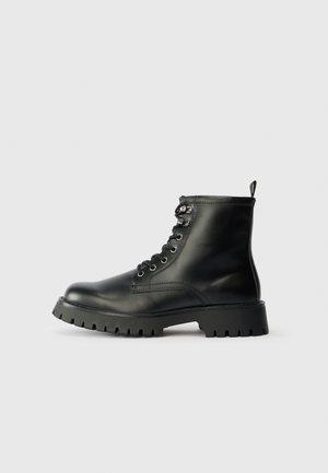 Stivaletti in pelle nera con punta rotonda, sei passanti per lacci e una suola spessa e texturizzata con scanalature profonde.