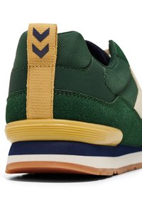 Green and beige sneaker with a textured upper, rubber sole, yellow heel tab, and navy accent. Features chevron detailing on the tab.
