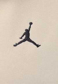Logo de Jumpman bordado en negro sobre un tejido beige texturizado, mostrando una figura en una pose de baloncesto en el aire contra un fondo claro.