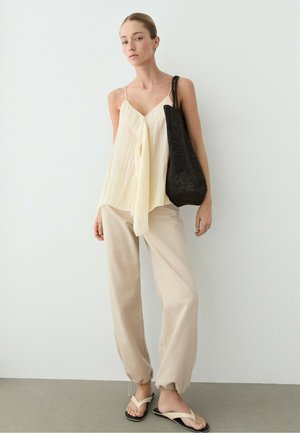Femme portant un haut crème ample, un pantalon beige avec des poignets à cordon et des sandales, portant un sac à bandoulière noir tissé, debout contre un mur blanc.