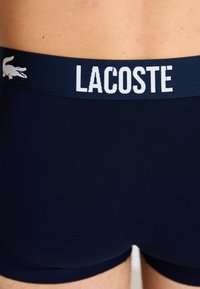 Tamsiai mėlynos spalvos medvilniniai bokseriai su balta "LACOSTE" logotipu elastingame dirželyje, pasižymintys sklandžia tekstūra ir prigludusia forma.