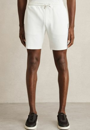 Witte, gestructureerde sportshorts met een elastische tailleband en trekkoord; met zwarte accenten aan de zijkanten. Gecombineerd met zwarte sneakers.