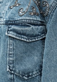 Τσέπη γαλάζιου τζιν με καπάκι και διακοσμητική κεντητή λεπτομέρεια με ασημένιες και διαφανείς χάντρες από πάνω.