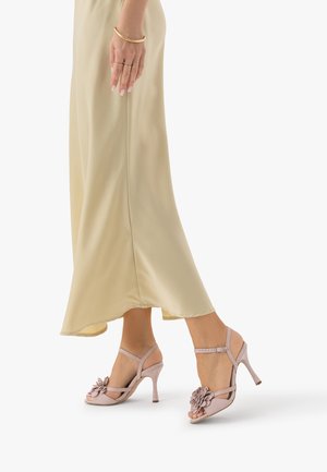 Kobieta w beżowej satynowej spódnicy i jasnoróżowych sandałach na wysokim obcasie z kwiatowymi zdobieniami robi krok do przodu na białym tle.