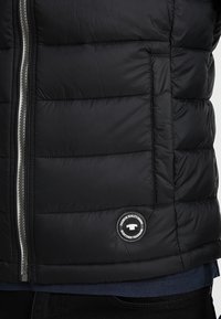 Schwarze Pufferjacke mit gestepptem Design, einem silbernen Reißverschluss, seitlichen Taschen und einem runden Logo-Patch auf der linken Seite.