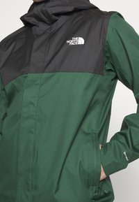 Veste imperméable verte et noire avec fermeture éclair devant, capuche et logo de la marque sur la poitrine. Texture lisse avec poches latérales.