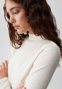 Cremefarbener, gerippter Rollkragenpullover mit dezenten schwarzen Punkten, langen Ärmeln und einer figurbetonten Silhouette. Weicher Materialtextur.