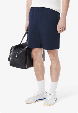 Navy blue elastic-waist shorts with side pockets, paired with white athletic socks and grey-blue sneakers, holding a black duffel bag.